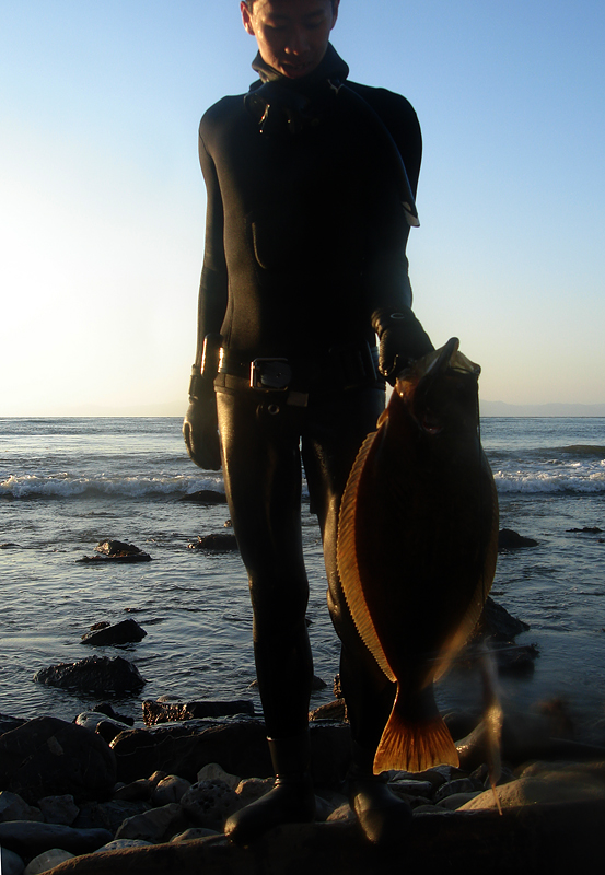 A man in a wetsuit stands on a rocky shore holding a small halibut