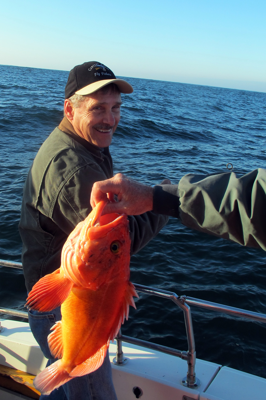 A man on a boat smiles as another man holds an orange rockfish for the camera