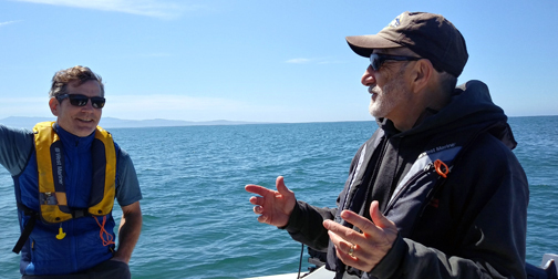 Two men stand talking on a boat