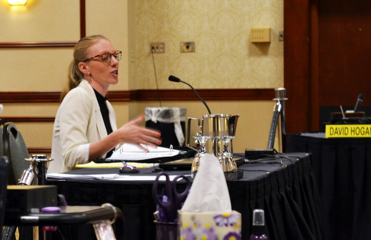 A woman wearing a white suit sits at a table talking into a microphone