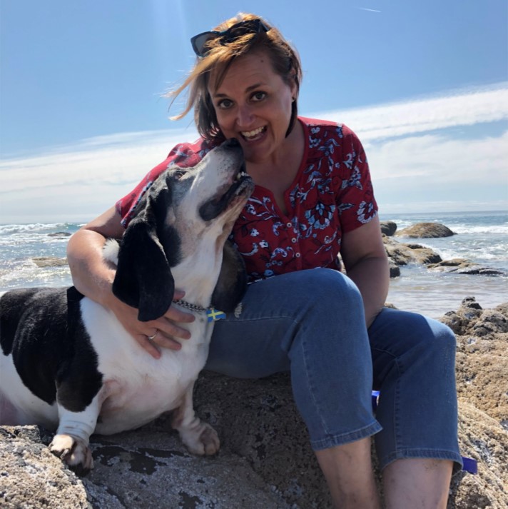 A woman in a red shirt sits on a rock at the beach, smiling with her dog