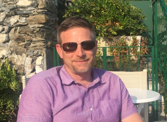 A man wearing sunglasses and a lavender shirt smiles for the camera
