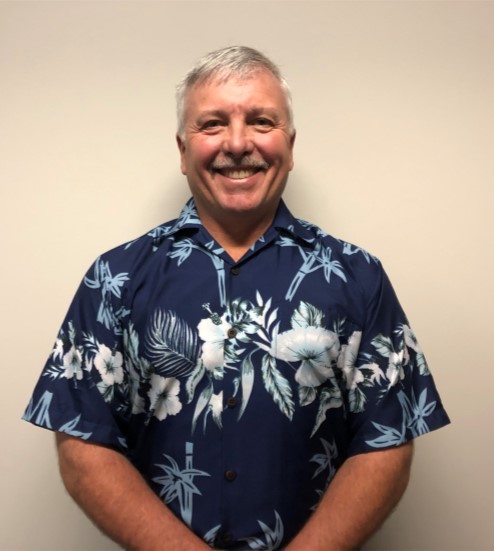 A man in a Hawaiian shirt stands against a wall, smiling