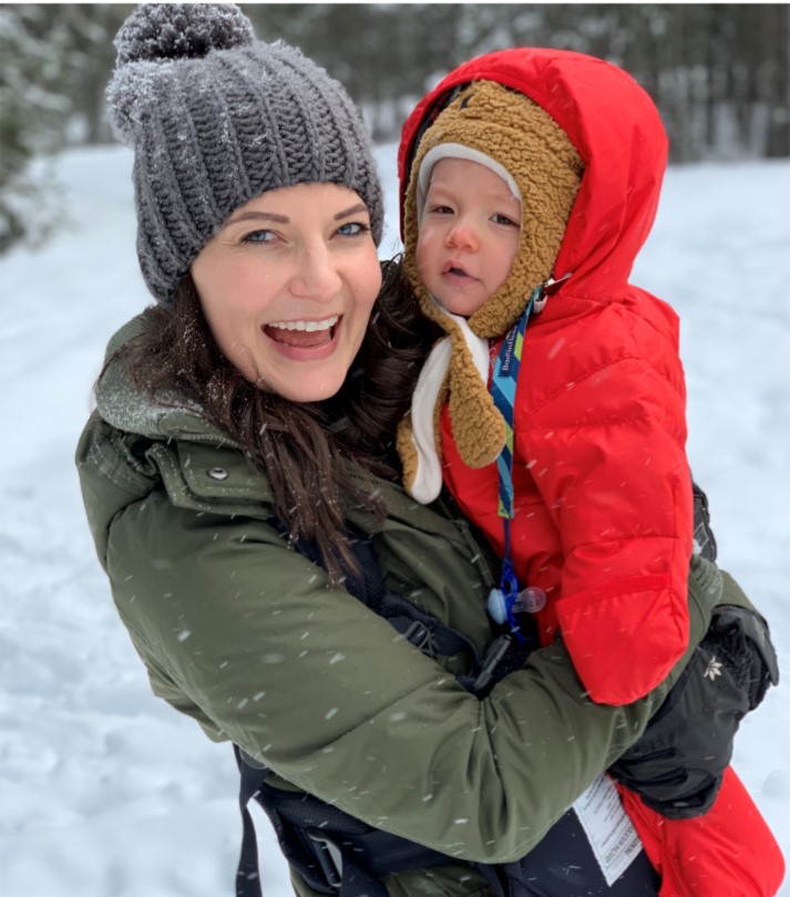 A woman in a knitted hat poses, smiling, with a baby in a the snow