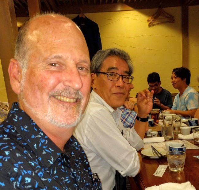 A man in a black Hawaiian shirt smiles at the camera