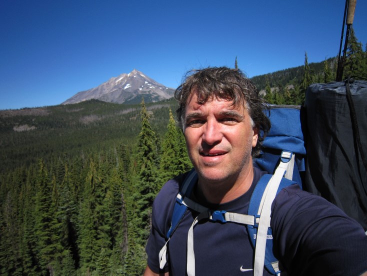 A man wearing a backpack poses in front of a mountain