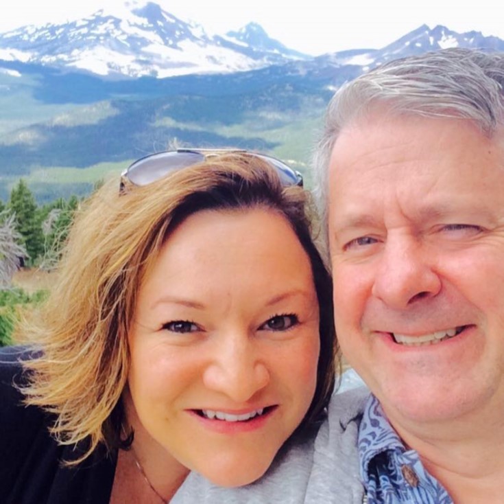 A woman and her husband pose smiling in front of a snow-capped mountain