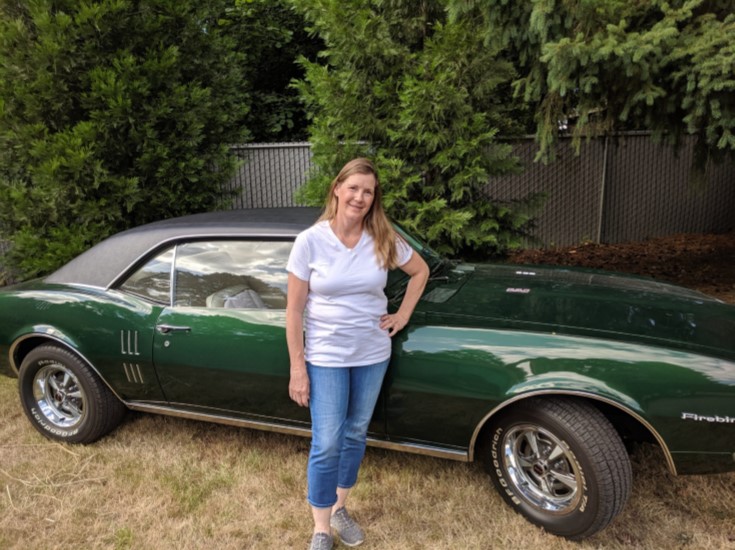 A woman in a white teeshirt and jeans stands smiling in front of a classic green sportscar